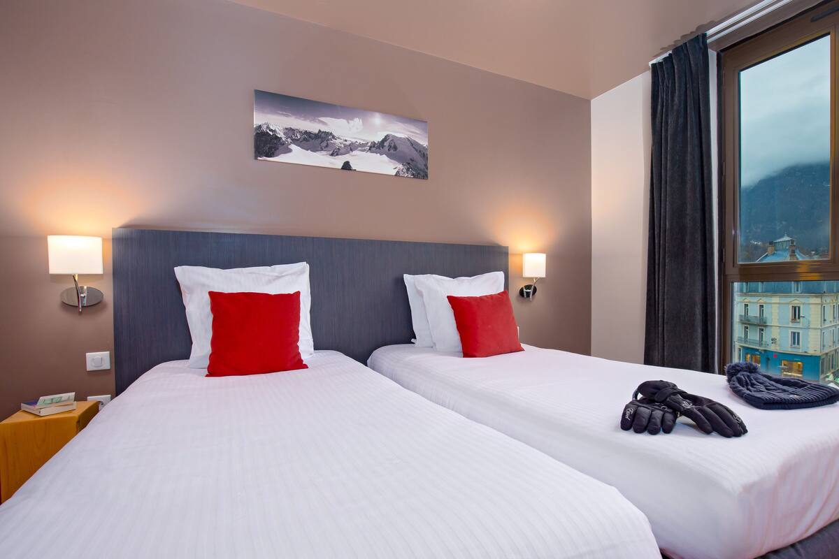 Ein Hotelzimmer mit zwei Betten, jeweils mit weißer Bettwäsche und roten Kissen. An der Wand hängt ein Gemälde von Bergen, an jedem Bett steht eine Lampe und das Fenster bietet Blick auf ein Gebäude.