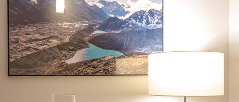 A wall-mounted picture of a mountainous landscape, a table with a candle and a tree-shaped ornament, and a lamp with a white shade.