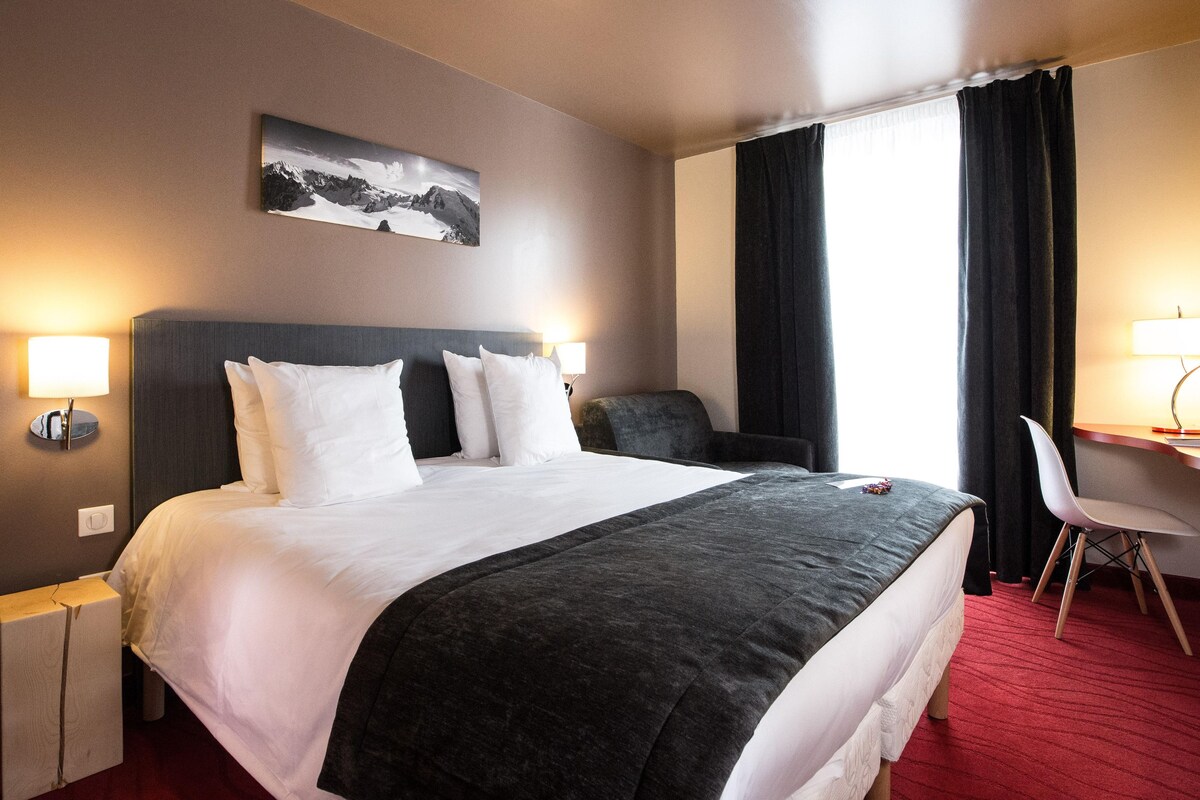 Et hotelværelse med en stor seng, to sengeborde med lamper, et skrivebord med stol, en sort stol og et billede på væggen.