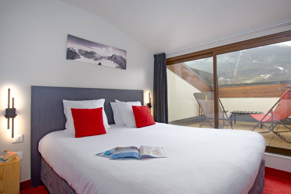 Ein Hotelzimmer mit einem großen Bett, roten und weißen Kissen, einem gerahmten Bild an der Wand und Blick ins Freie durch ein großes Fenster.