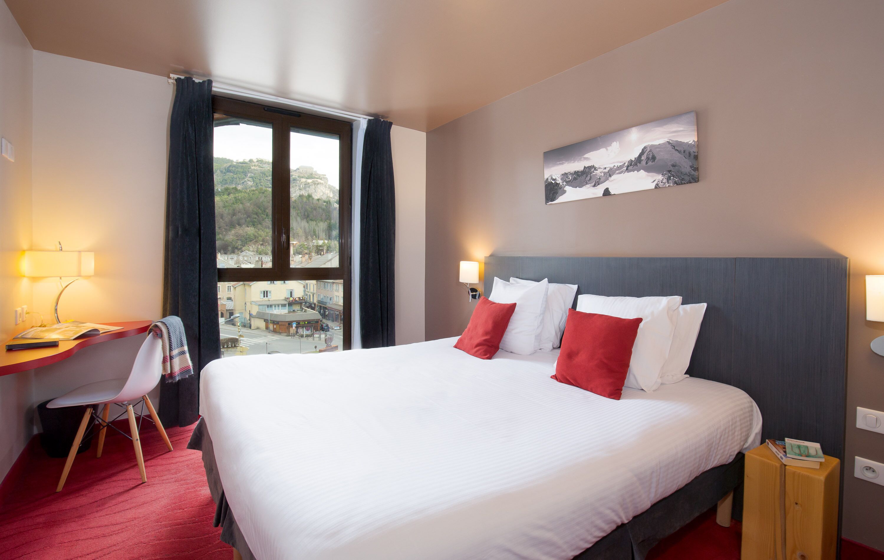 A hotel room with a large bed, a desk with a chair, a window with curtains, and a painting on the wall.