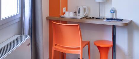 A room with a wooden desk, an orange chair, a small orange stool, a white kettle, a lamp, and a water bottle.
