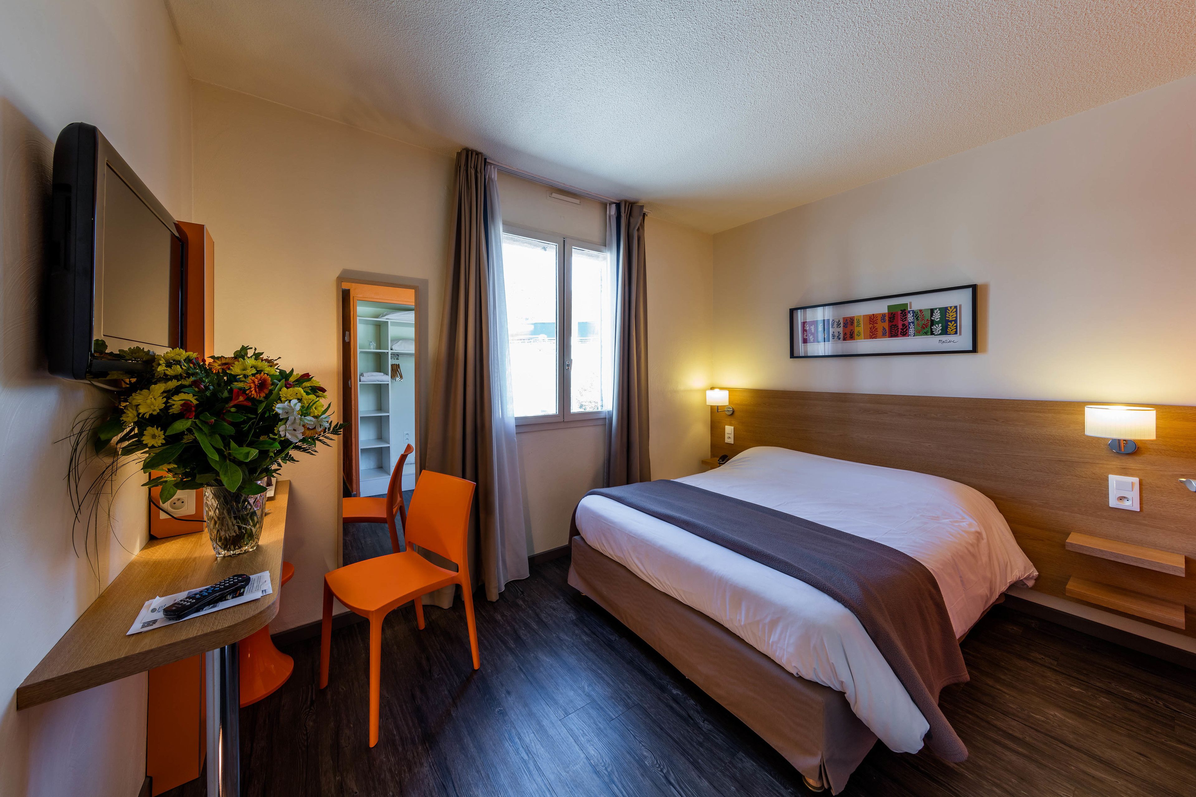 Ein Hotelzimmer mit einem Bett, einem Schreibtisch mit Stuhl, einem Fernseher, einer Vase mit Blumen, einem Fenster mit Vorhängen und einem Wandbild.