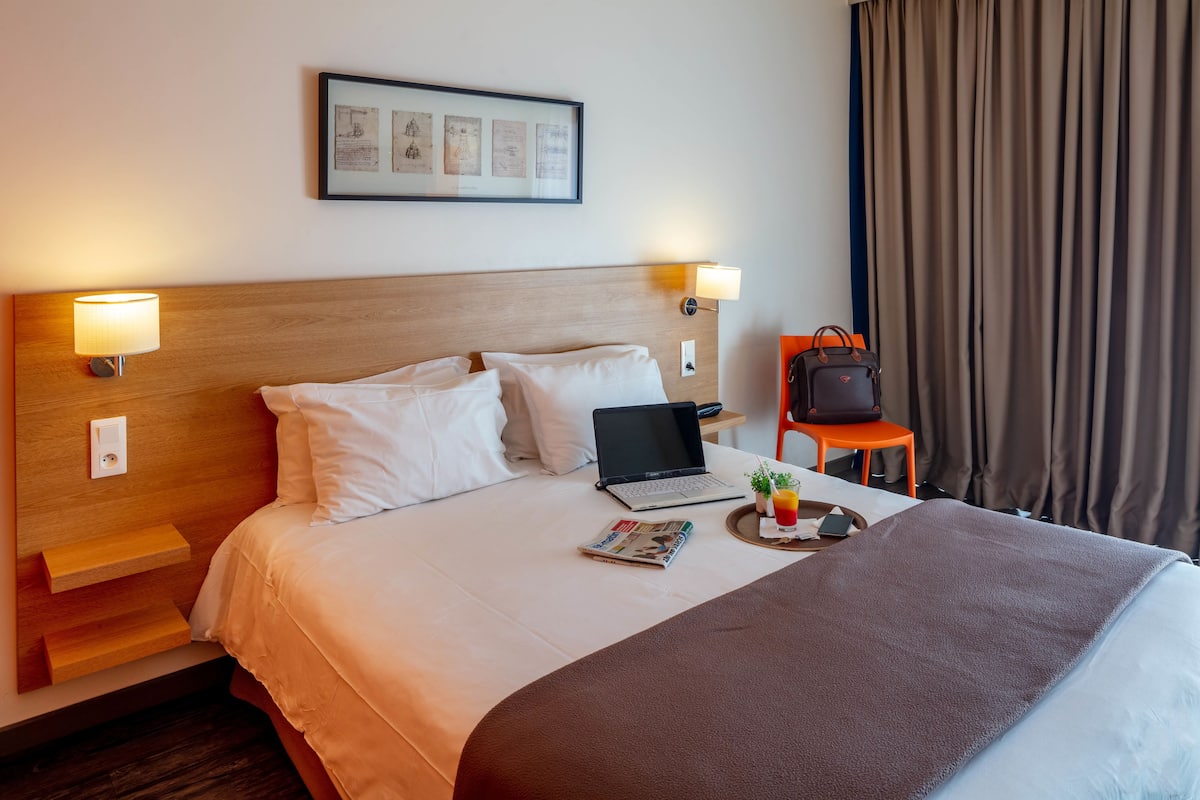Un lit bien fait, avec un ordinateur portable, un plateau contenant une boisson et un livre, et un sac à main posé sur une petite table à côté du lit.