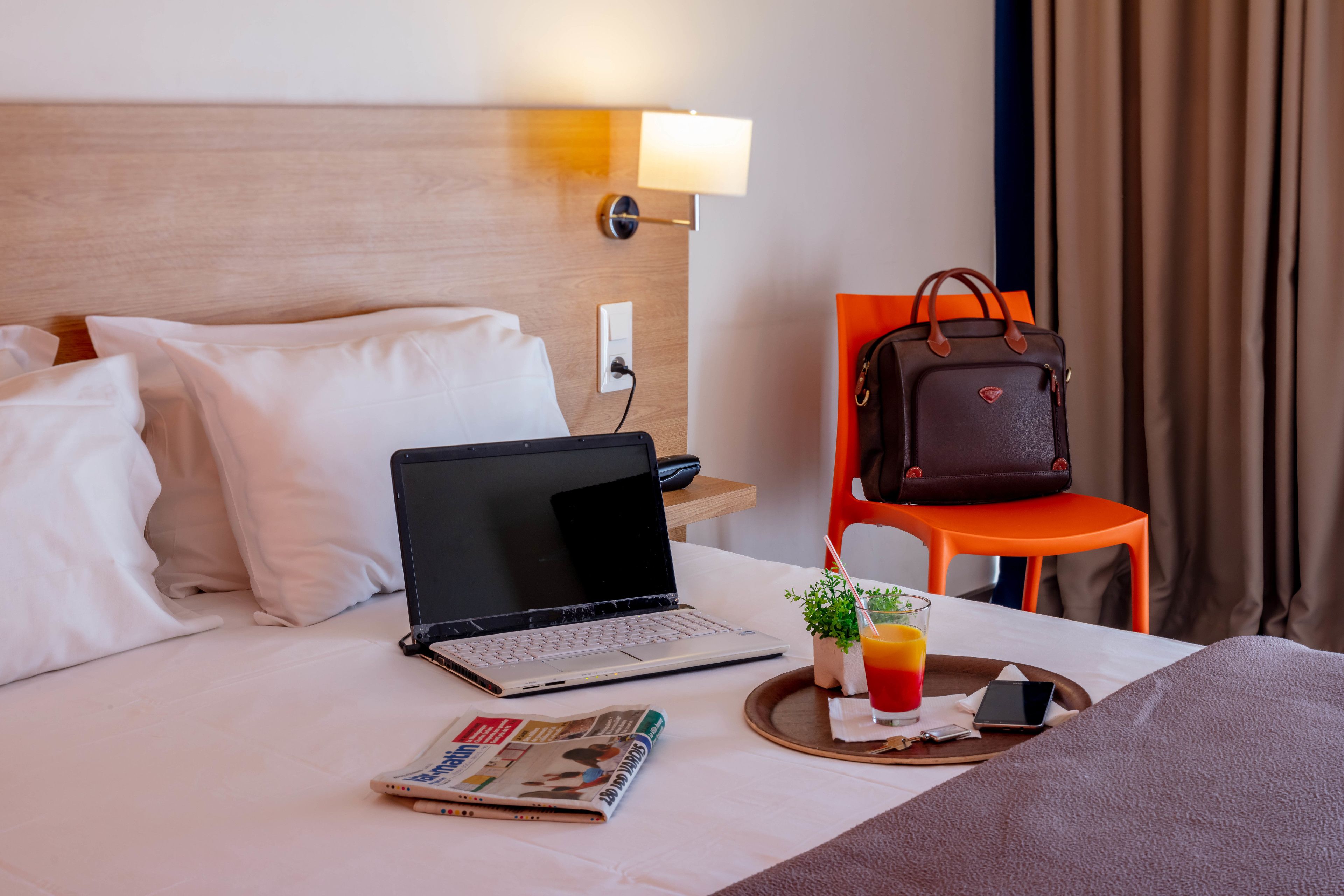 Una habitación de hotel impecable con cama, una laptop, una revista, una bandeja con una bebida y un bolso sobre una silla.