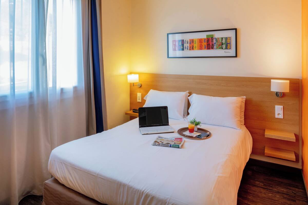 Un lit bien fait, avec du linge de lit blanc, un ordinateur portable, un plateau avec une boisson et un livre, et une estampe botanique encadrée accrochée au mur.