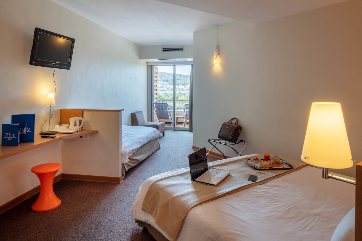 Une chambre d’hôtel avec un lit, un bureau avec un ordinateur portable, une télévision, une chaise, une table avec un petit-déjeuner, et un balcon avec vue.