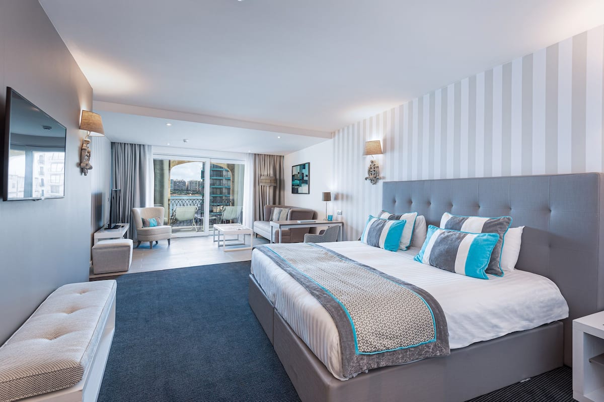 Ein modernes Hotelzimmer mit einem großen Bett, einer Sitzecke mit einer Couch und Sesseln, einem Esstisch und einem Balkon mit Blick auf die Stadt.