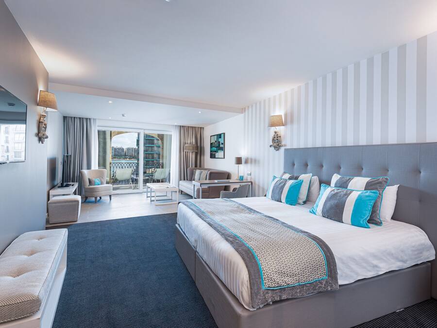 A modern hotel room with a large bed, a seating area with a sofa and armchairs, a dining table, and a balcony with a view of the city.