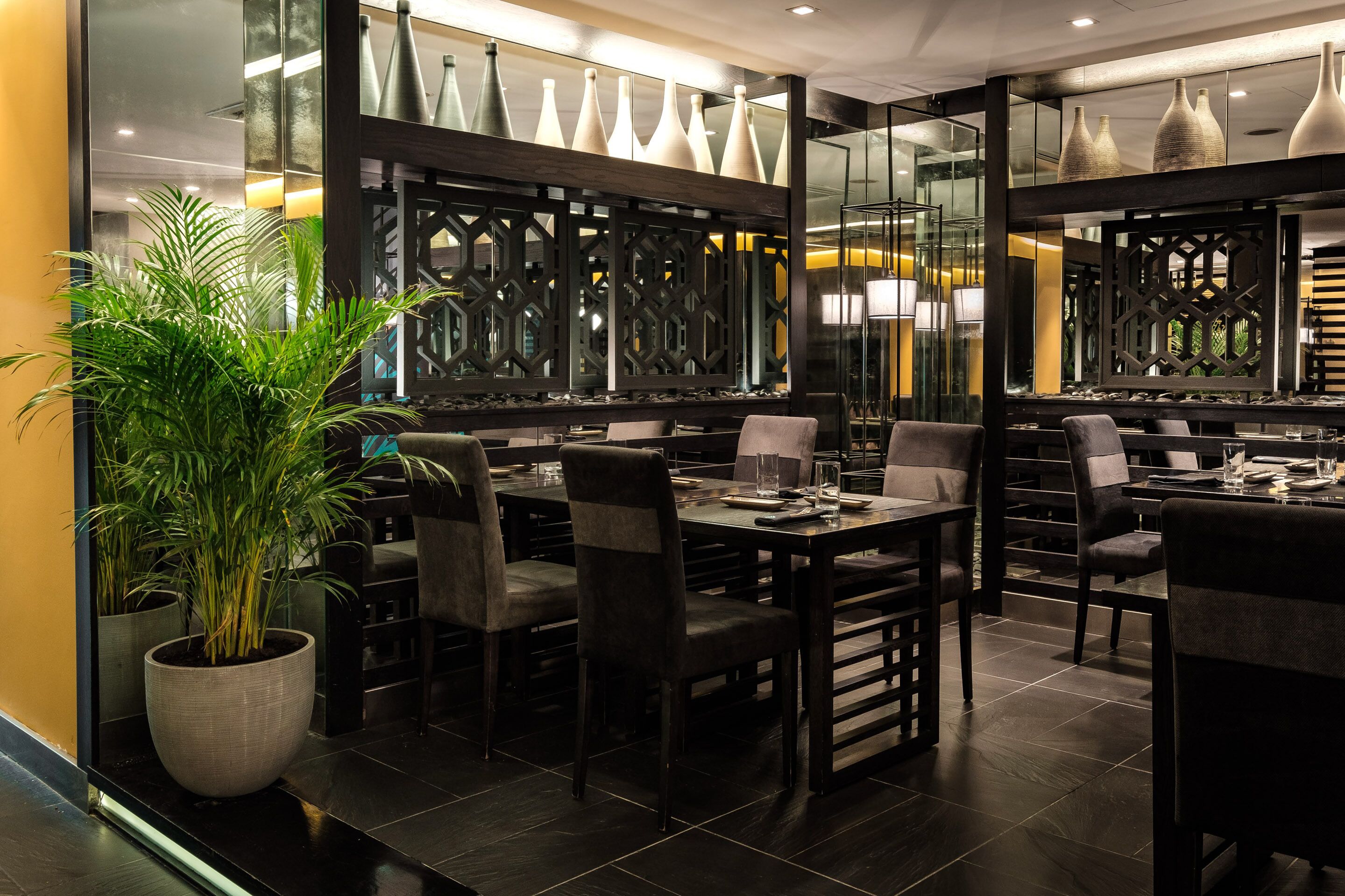 Interior de restaurante moderno com móveis de madeira escura, uma grande planta em vaso e vasos decorativos.