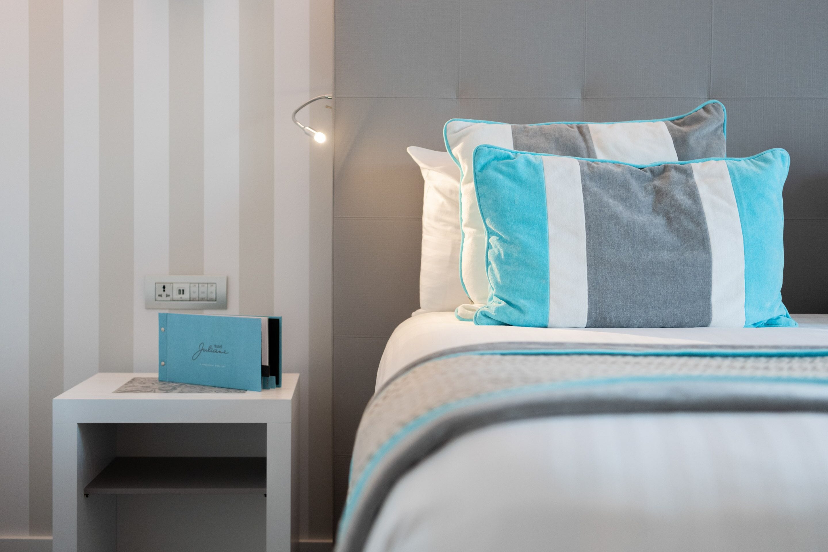 A neatly made bed with striped pillows, a bedside table with a blue folder, and a wall with vertical stripes.