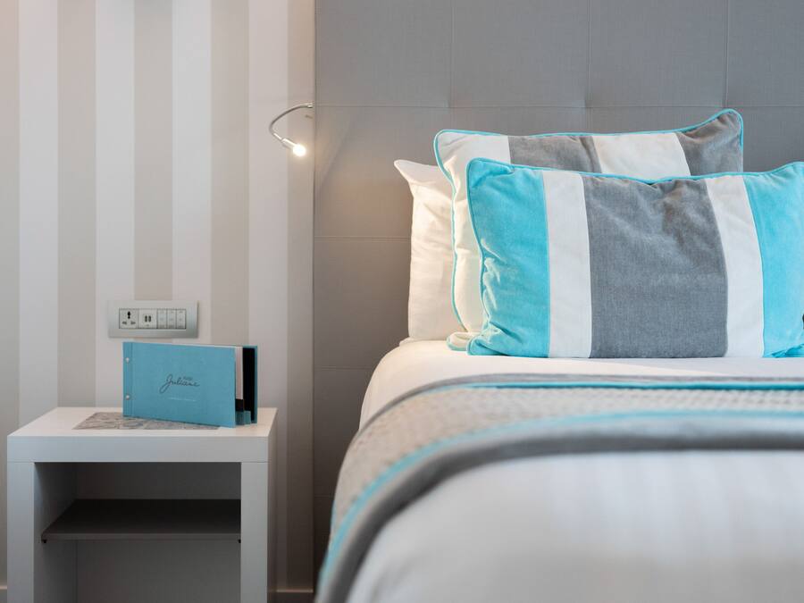 Una cama bien hecha con almohadones a rayas, una mesita de noche con una carpeta azul, y una pared con rayas verticales.