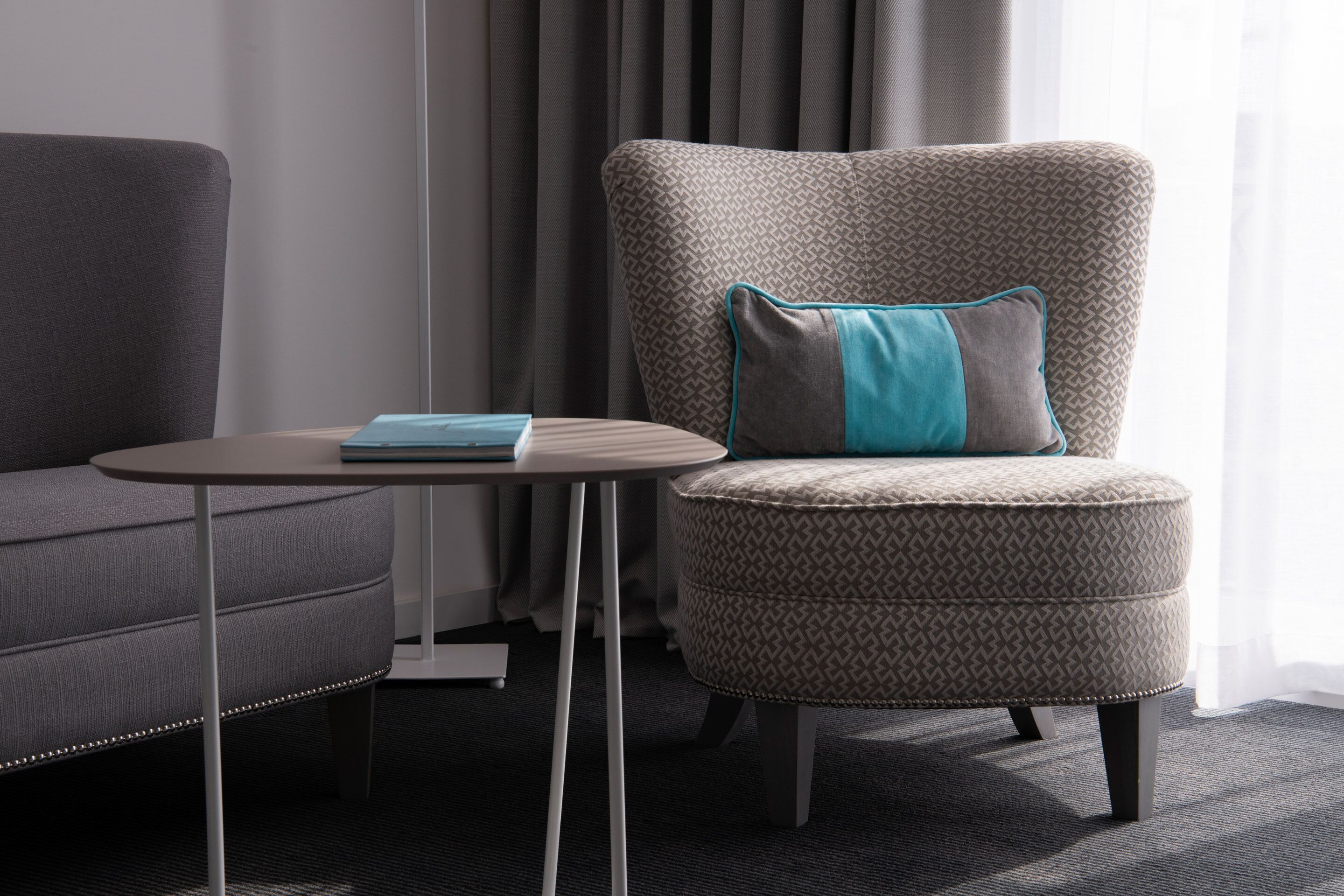 A modern room with a grey sofa, a patterned armchair with a teal pillow, and a small round table with a book.