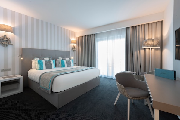 A modern hotel room with a large bed, bedside lamps, a desk with a chair, and a view of the outdoors through sheer curtains. - Hotel Juliani (St. Julian's)