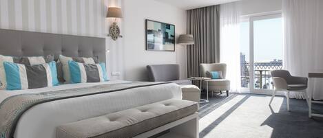 A modern hotel room with a large bed, a seating area with a sofa and armchairs, a small table, and a balcony with a view.