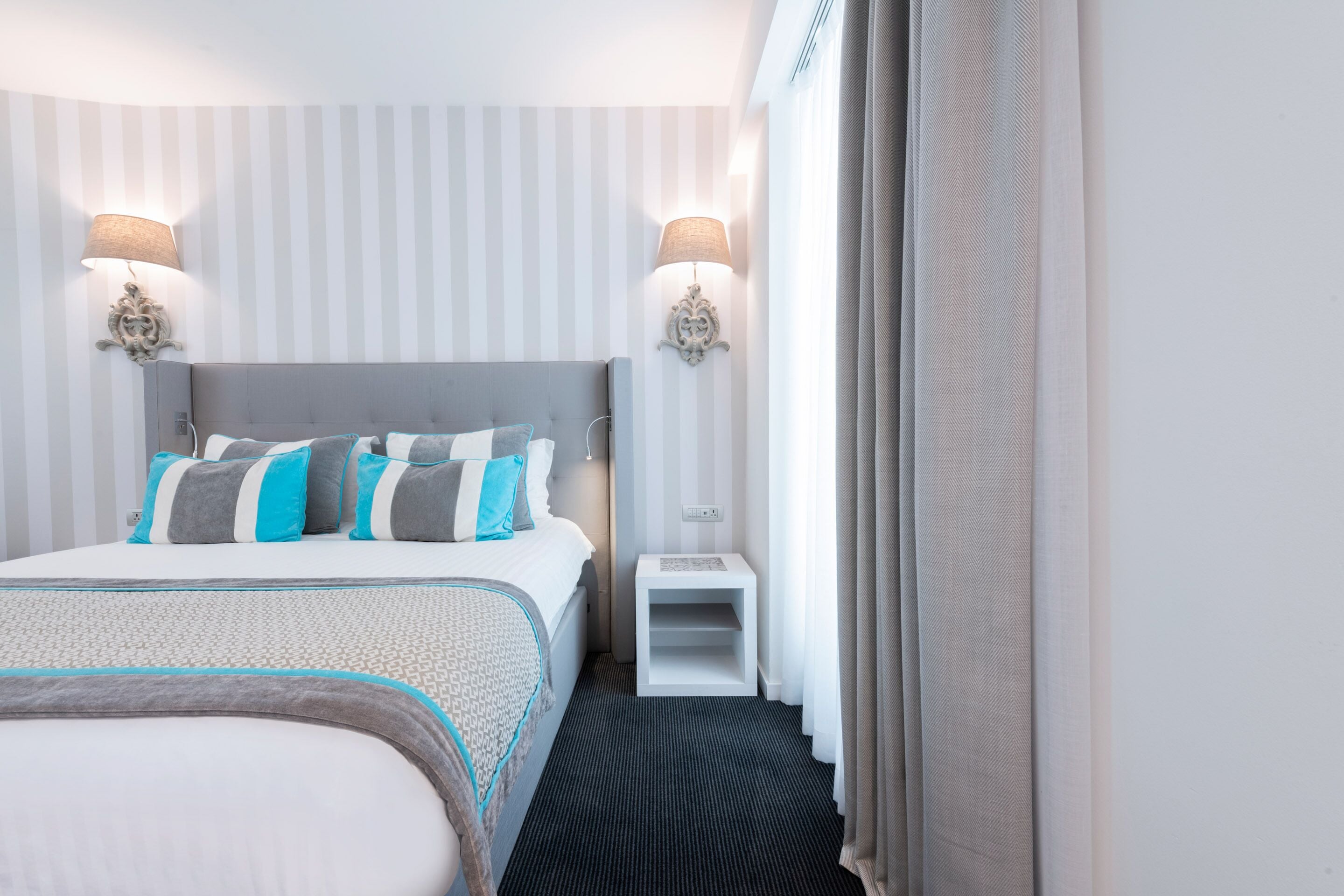 A hotel room with a large bed, two bedside lamps, a small nightstand, and a window with curtains.