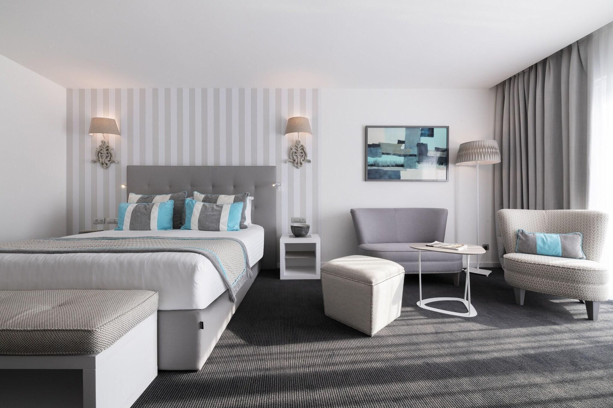 A modern hotel room with a large bed, two bedside lamps, a gray armchair, a small side table, and a gray sofa.