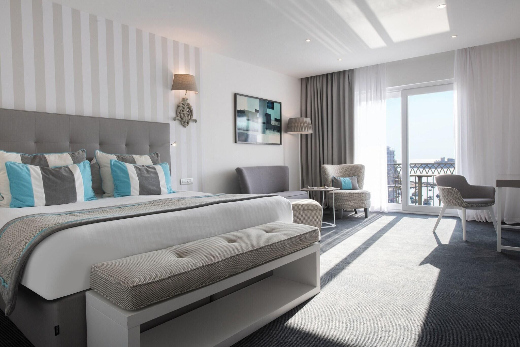 A modern hotel room with a large bed, a seating area, a balcony, and a city view.