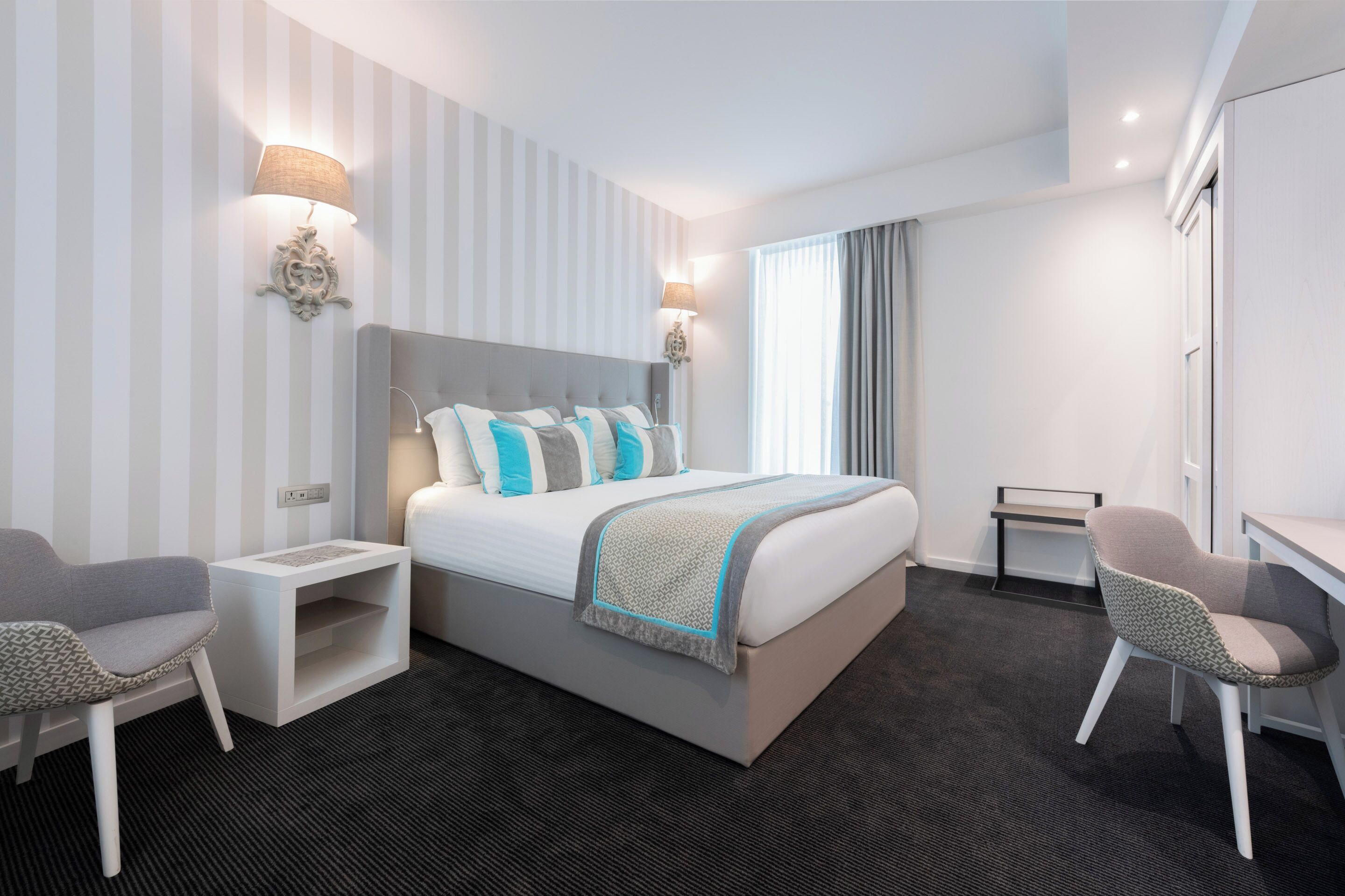 A hotel room with a bed, bedside tables, a chair, a desk, and a window with curtains.