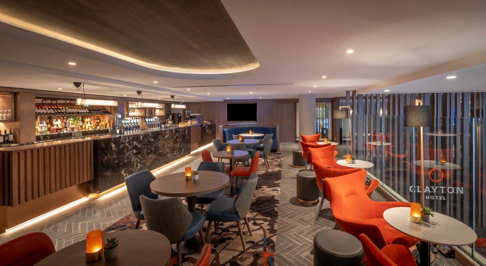 A modern hotel bar with a bar counter, seating area with round tables and orange chairs, and a visible television screen.
