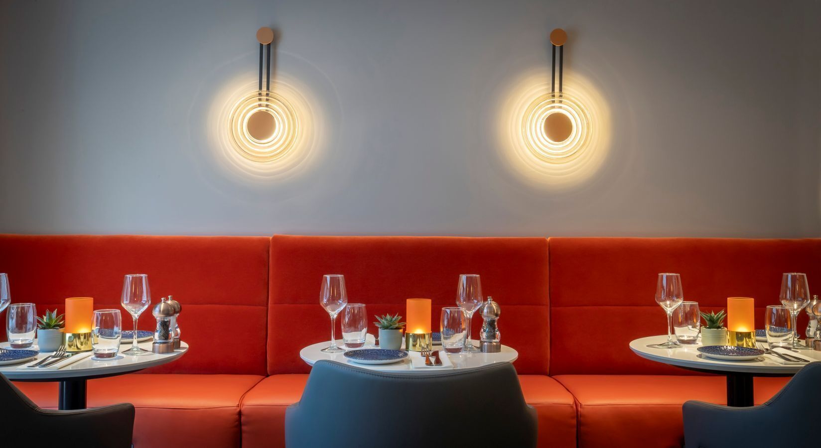 Un restaurante con asientos tapizados en rojo, mesas redondas con manteles blancos y dos lámparas fijadas a la pared.