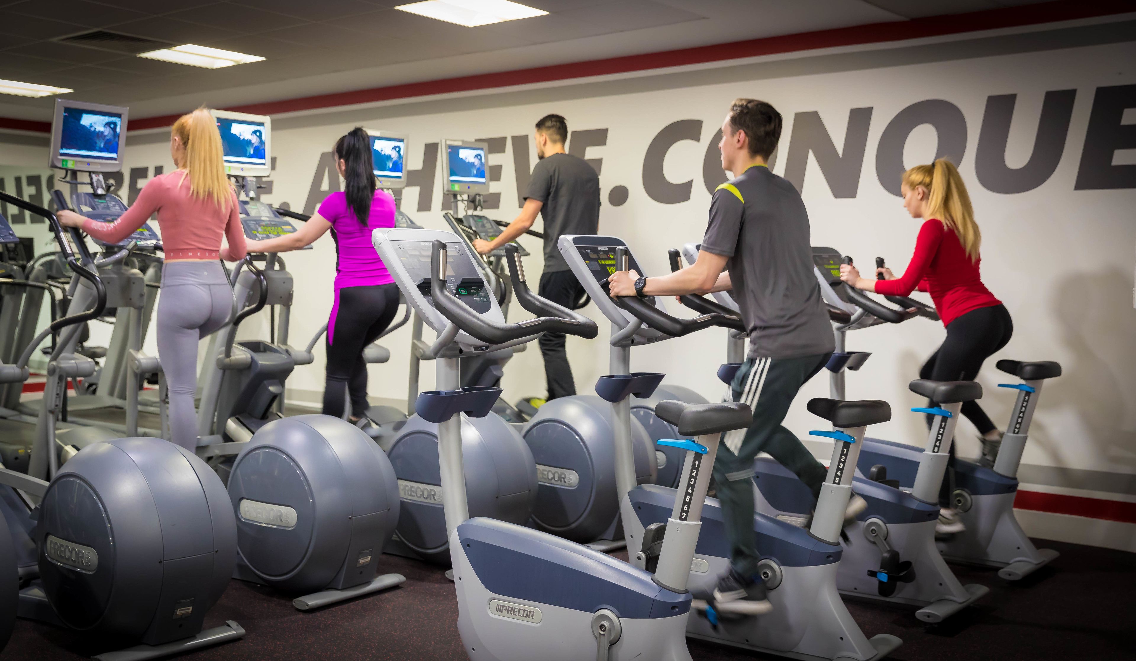 Un gimnasio con varias cintas de correr y bicicletas estáticas, un espejo grande y carteles de motivación en la pared.