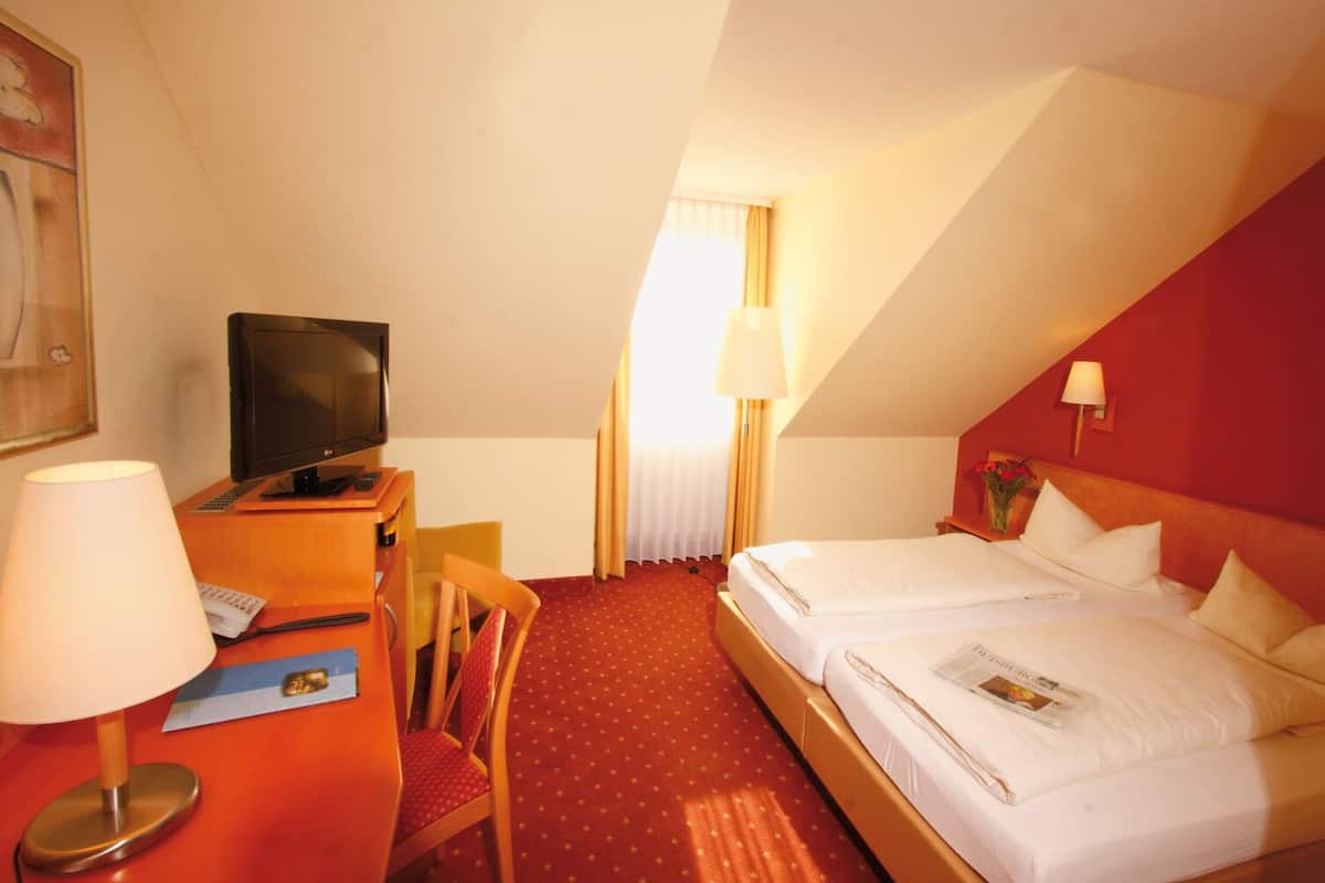 Ein Hotelzimmer mit Schreibtisch, Stuhl, Fernseher, zwei Betten und einem Fenster mit Vorhängen.