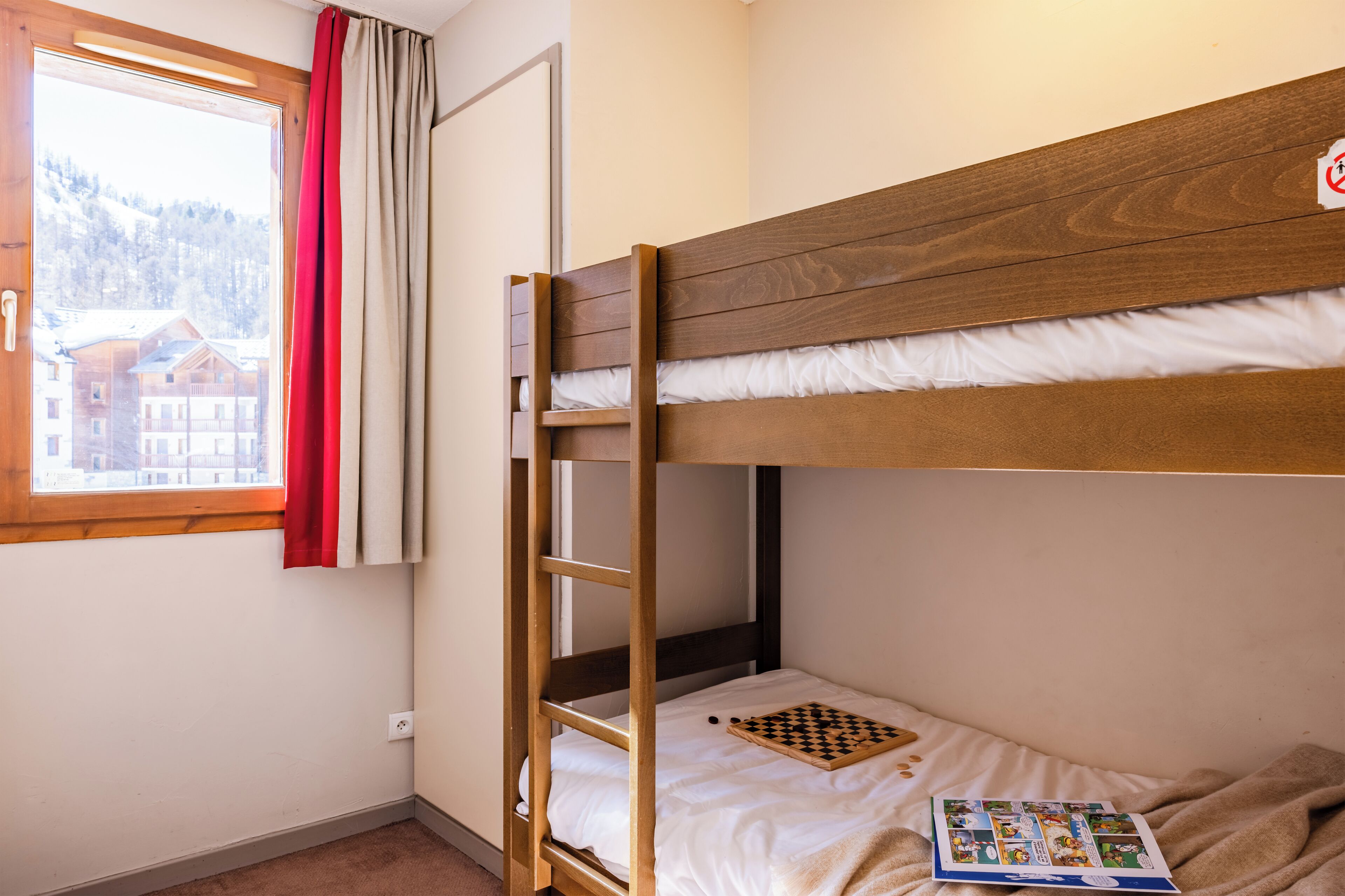 A bunk bed room with a window, a red curtain, and a chessboard on the lower bunk.