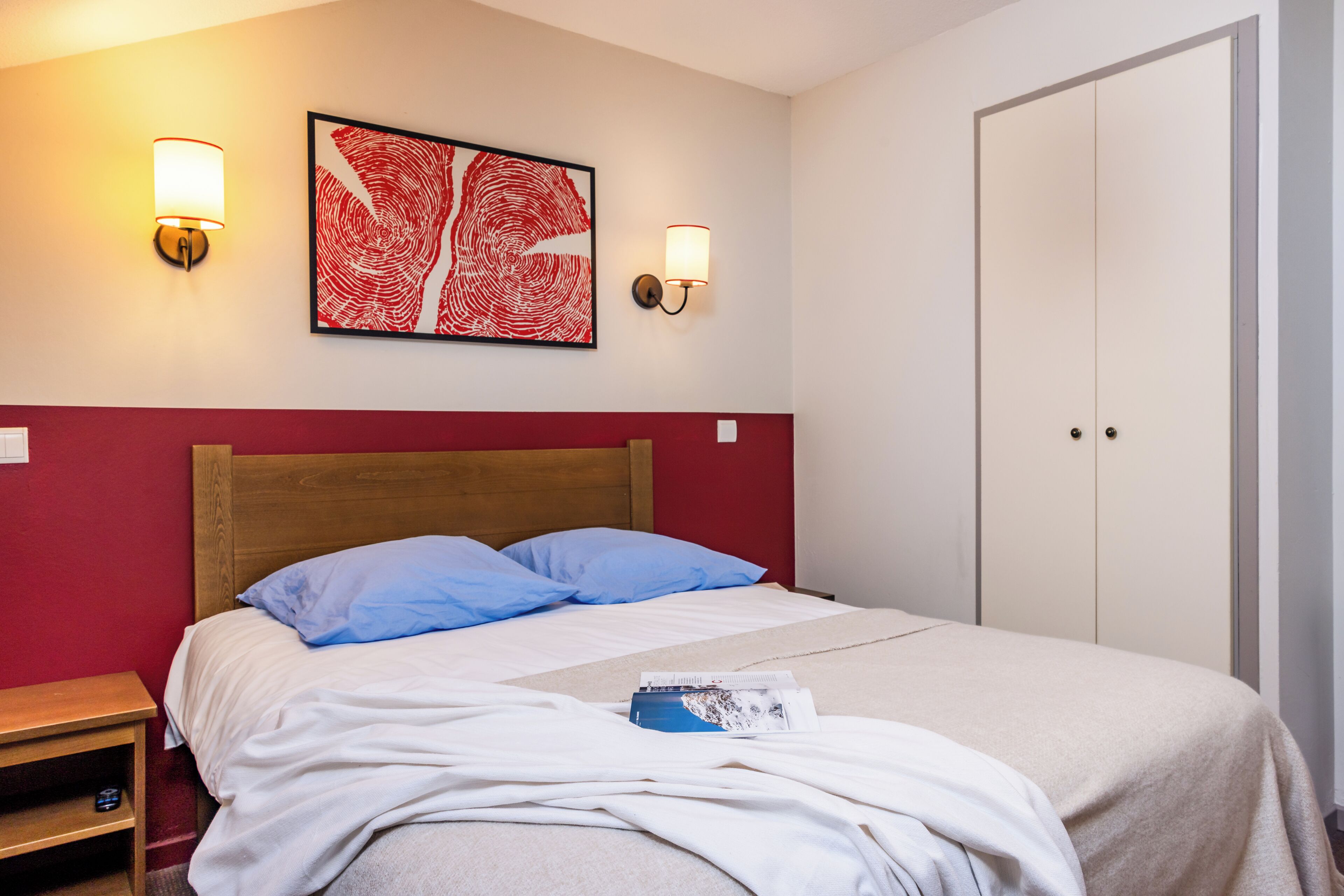 A neatly made bed with blue pillows, a red wall, and a framed artwork above it.
