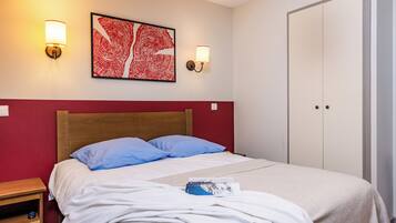 A neatly made bed with blue pillows, a red wall, and a framed artwork above it.