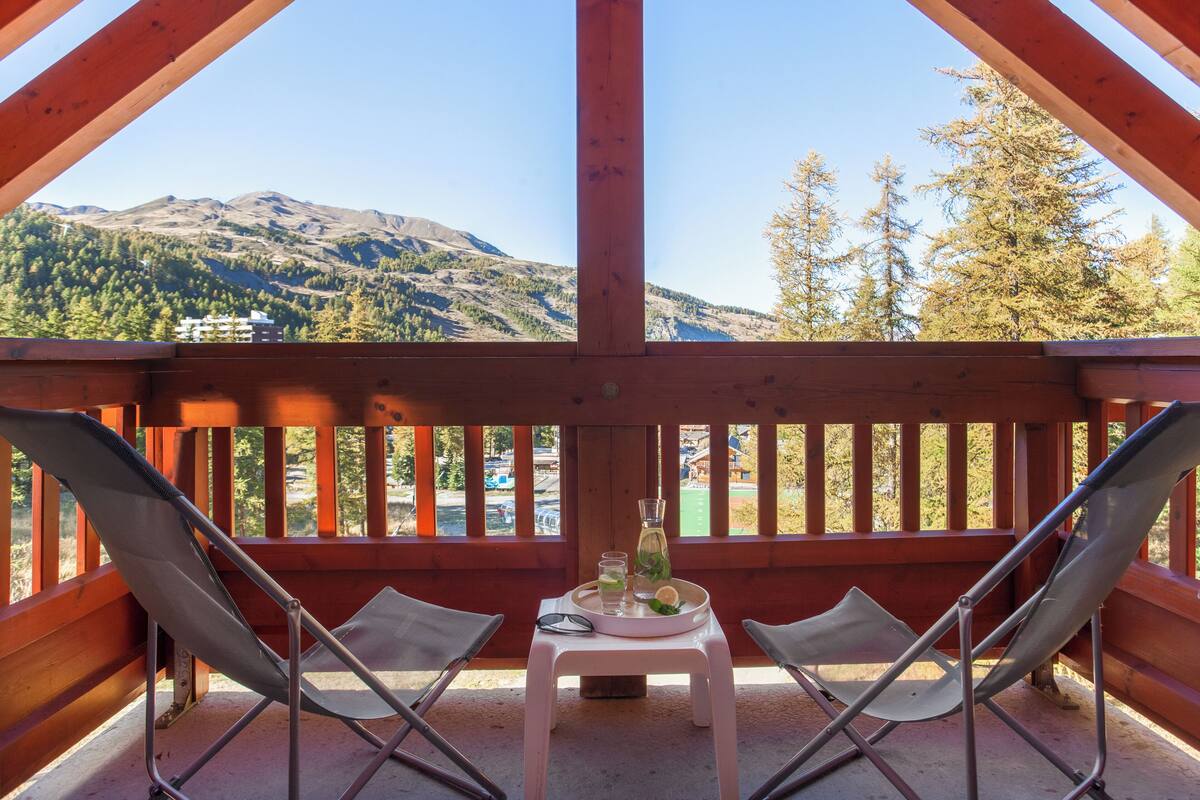 A wooden deck with two metal chairs, a small table with a bottle and glasses, and a view of mountains and trees.