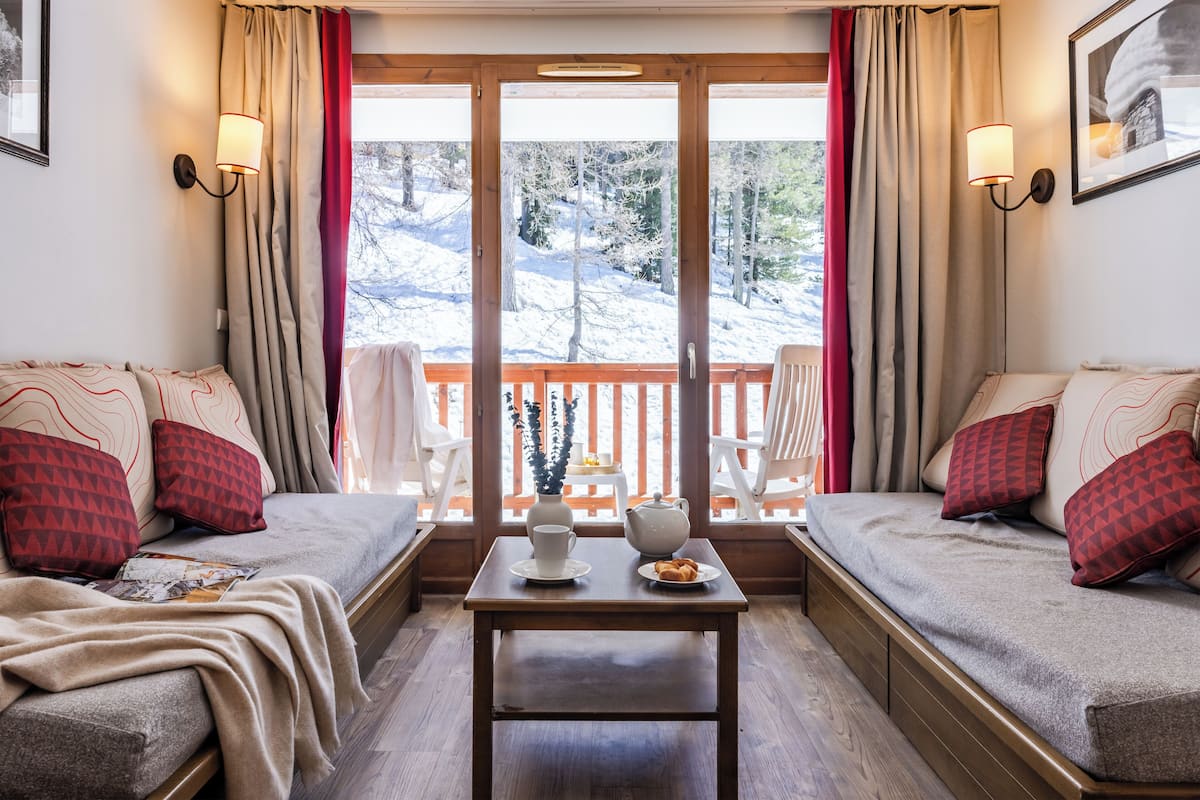 A cozy room with a sofa, a small table with a cup and a vase, and a view of a snowy landscape through a glass door.