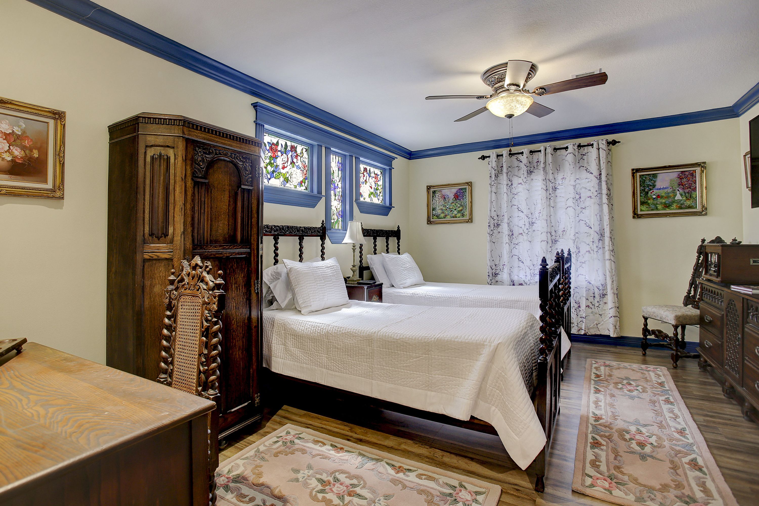 Une chambre avec deux lits, une armoire en bois, un ventilateur de plafond et un tapis à motifs floraux.