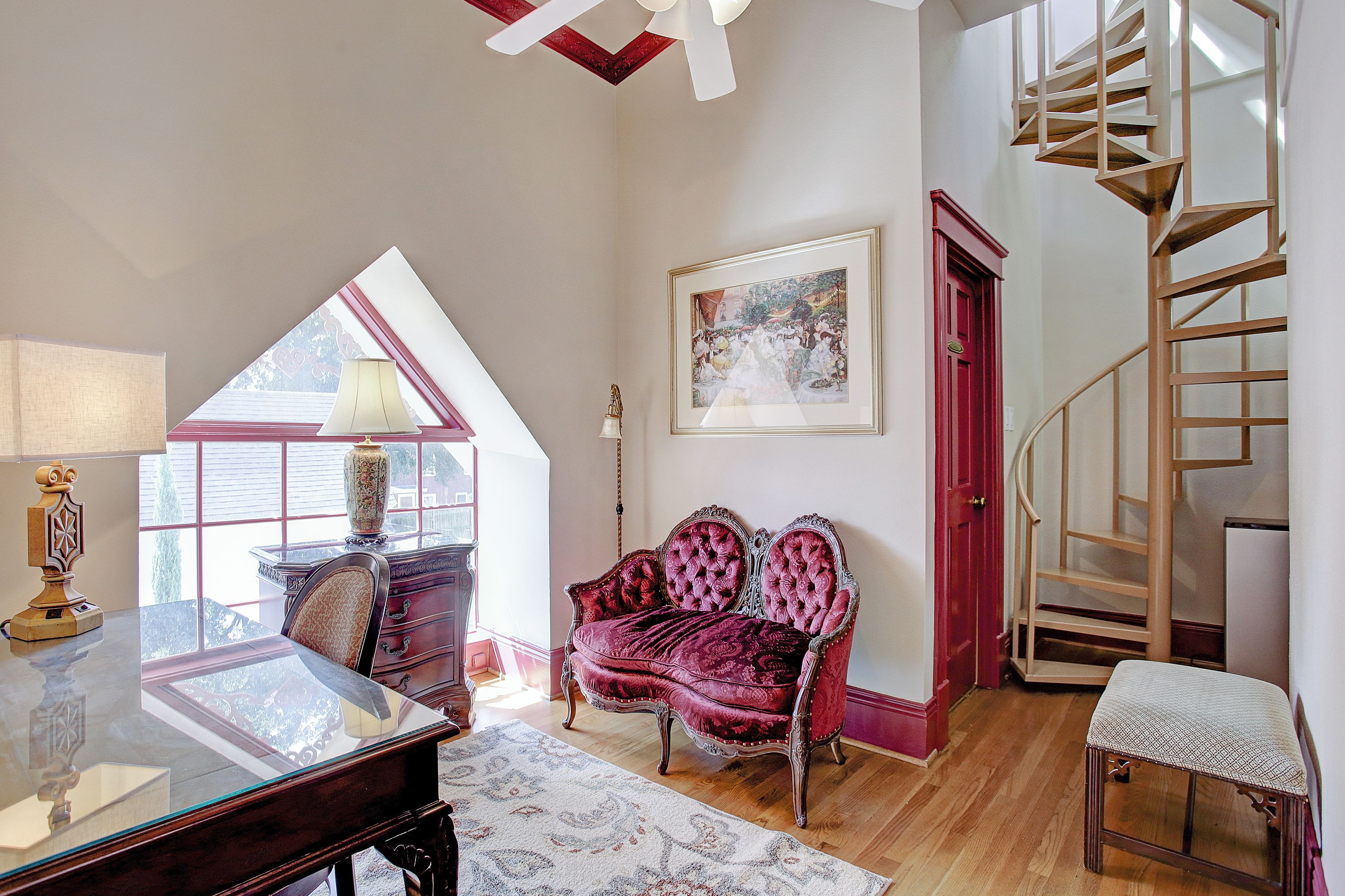 A spacious room with a glass desk, a red velvet sofa, a wooden chair, a small table, a framed painting, and a spiral staircase.