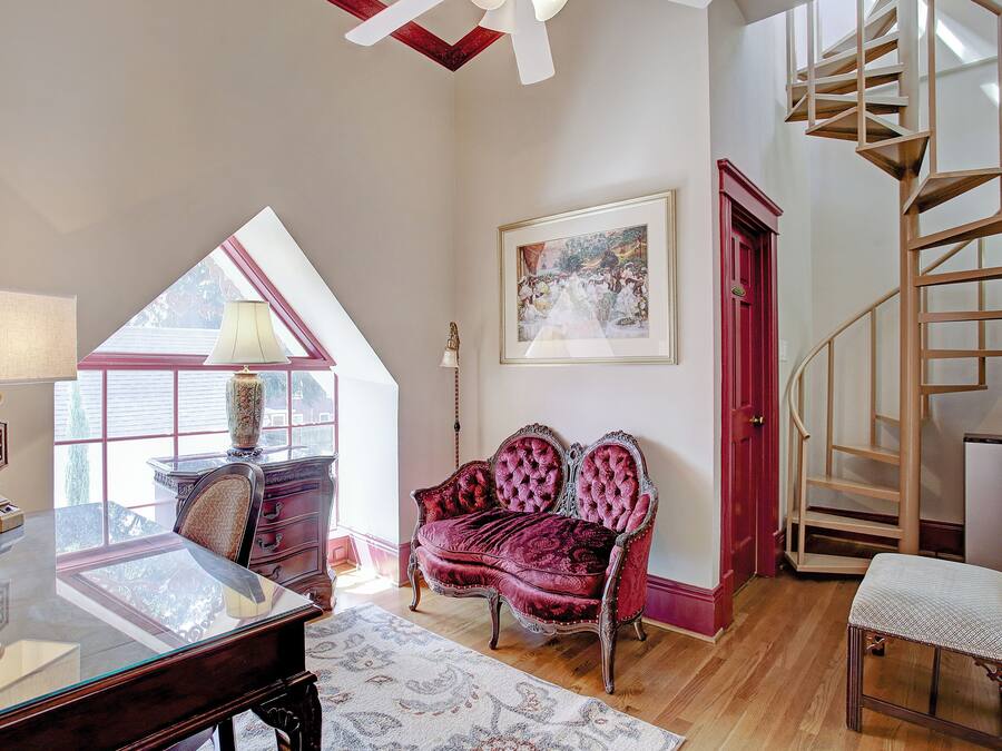 A spacious room with a glass desk, a red velvet sofa, a wooden chair, a small table, a framed painting, and a spiral staircase.