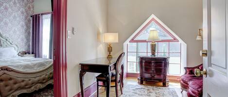 A bedroom with a large bed, a desk, and a dresser. There is a window with a view of the outside.