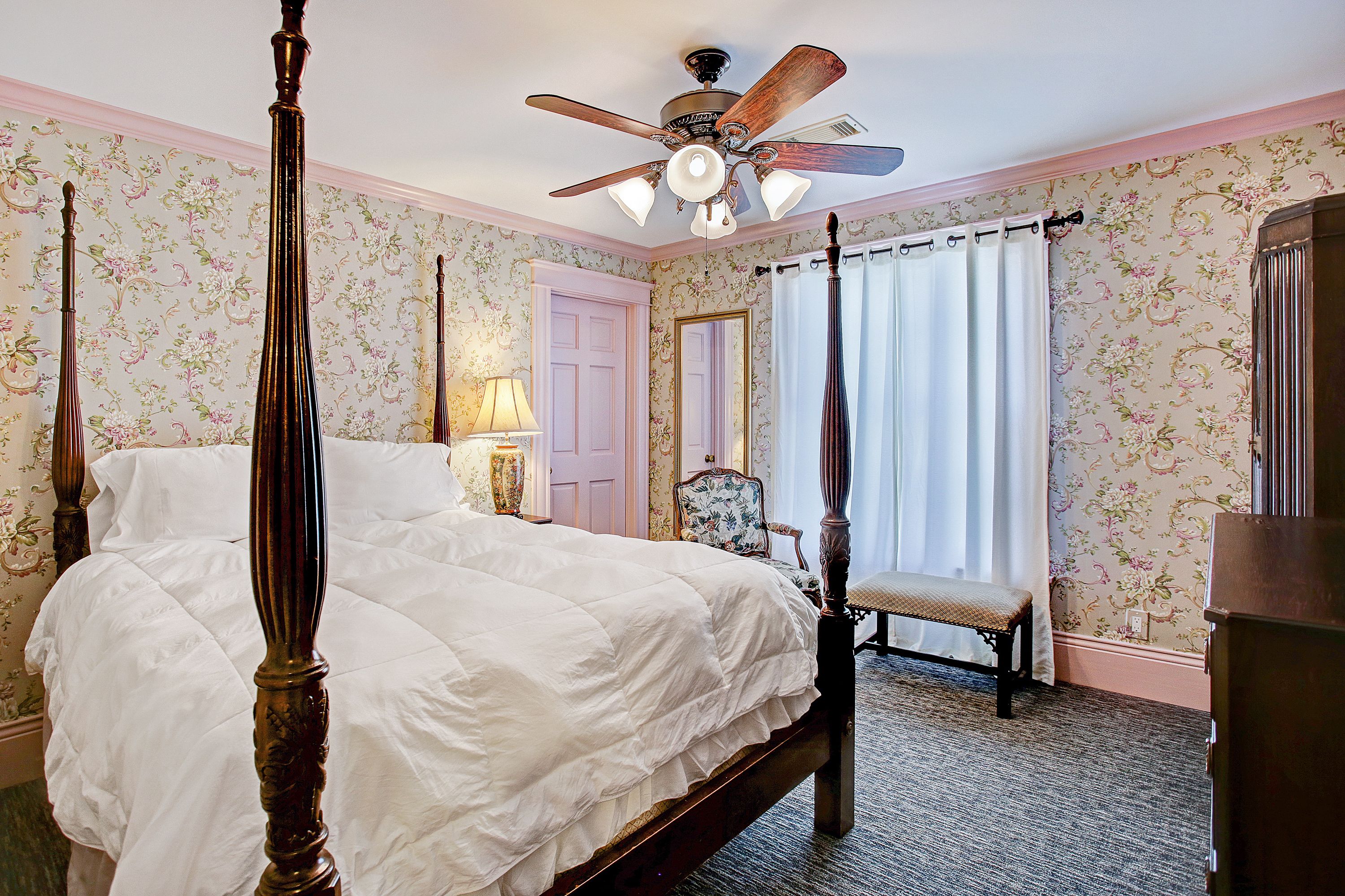 A four-poster bed with a ceiling fan, floral wallpaper, and a bench by the window.