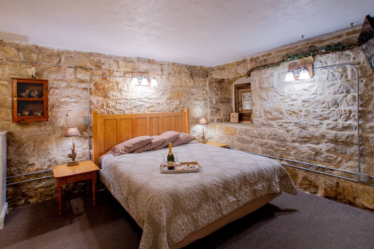 A bedroom with a stone wall, a wooden bed, a nightstand with a lamp, and a dresser.