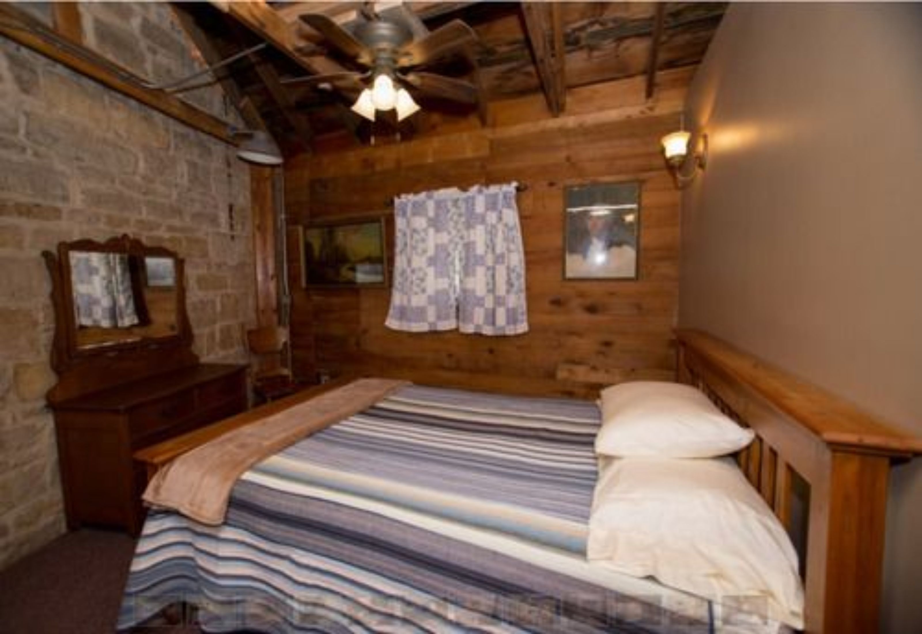 Un dormitorio de cabaña de madera con una cama, un ventilador de techo, una ventana con cortinas y un aparador de madera.
