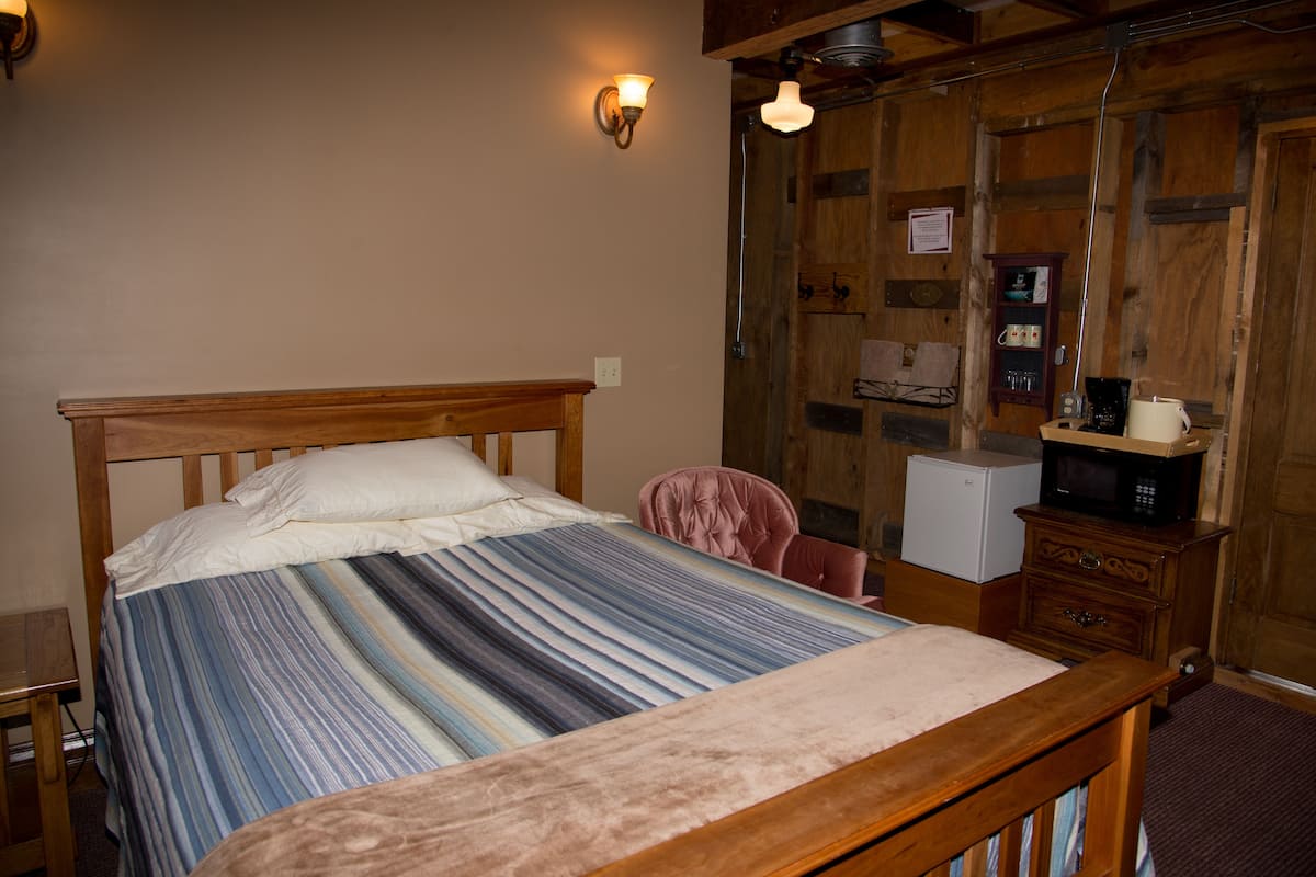 A bedroom with a wooden bed, a small desk, a chair, and a microwave on a wooden cabinet.