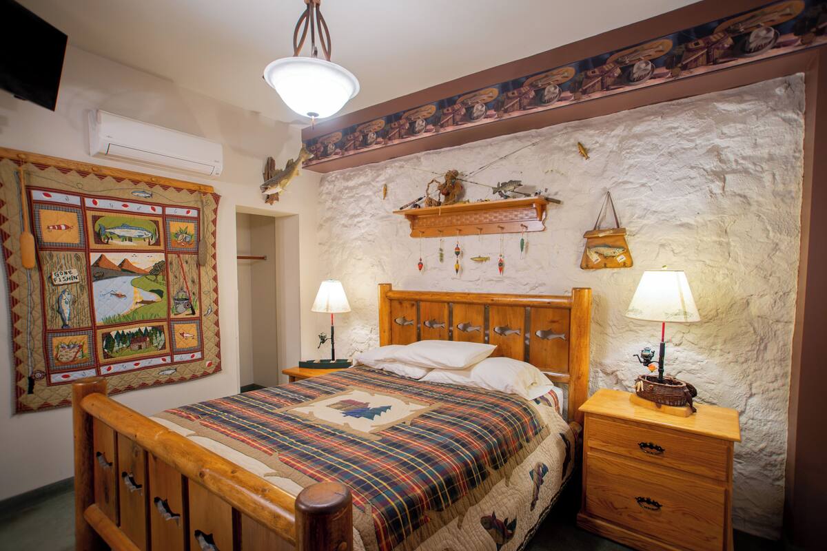 A bedroom with a wooden bed, two bedside tables with lamps, a quilt with fish patterns, a wall tapestry, and a stone wall.