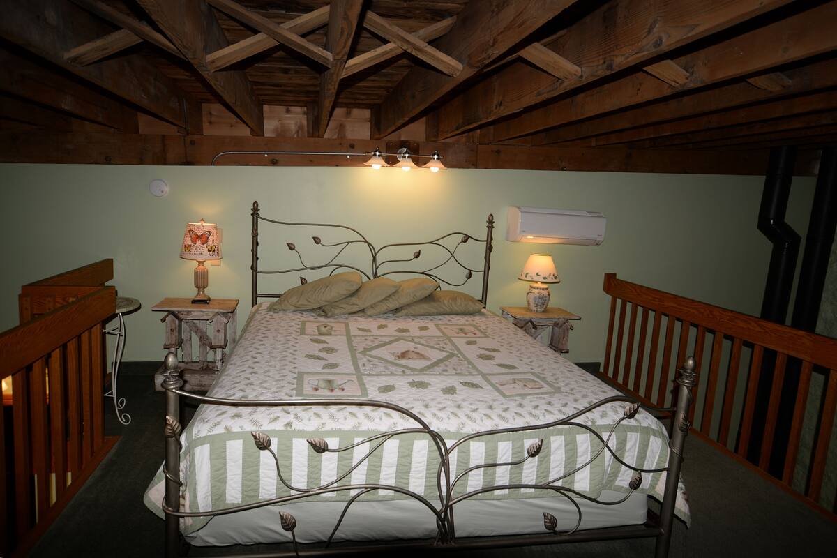 A bedroom with a wooden ceiling, a metal bed frame, two bedside tables with lamps, and an air conditioning unit.