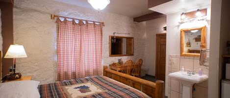 Een slaapkamer met een bed, nachtkastje, kapstok en badkamer met een wastafel en spiegel.