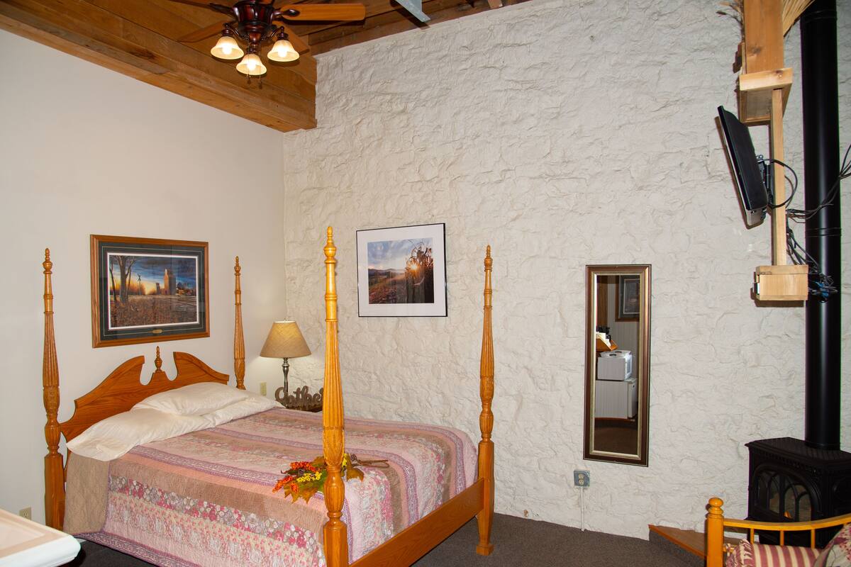 A bedroom with a four-poster bed, a wooden ceiling, a stone wall, a framed picture, a lamp, and a television mounted on the wall.