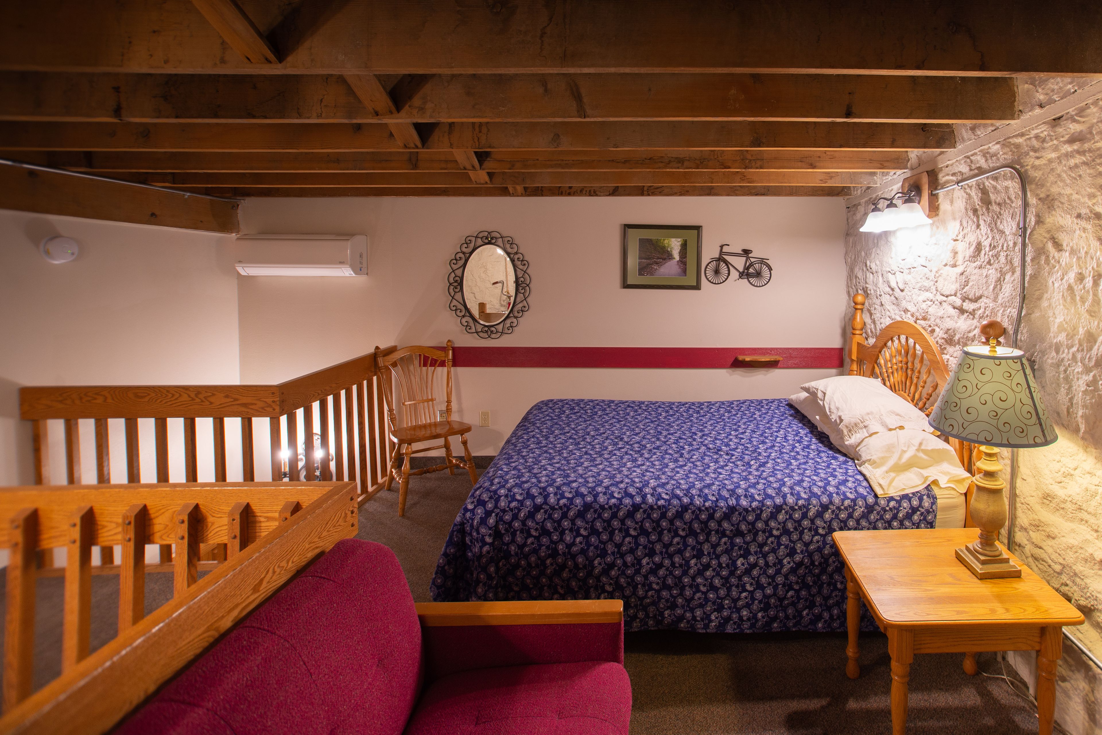 Un dormitorio con escalera de madera, cama con edredón estampado, una silla, una mesa con lámpara y un espejo fijado a la pared.