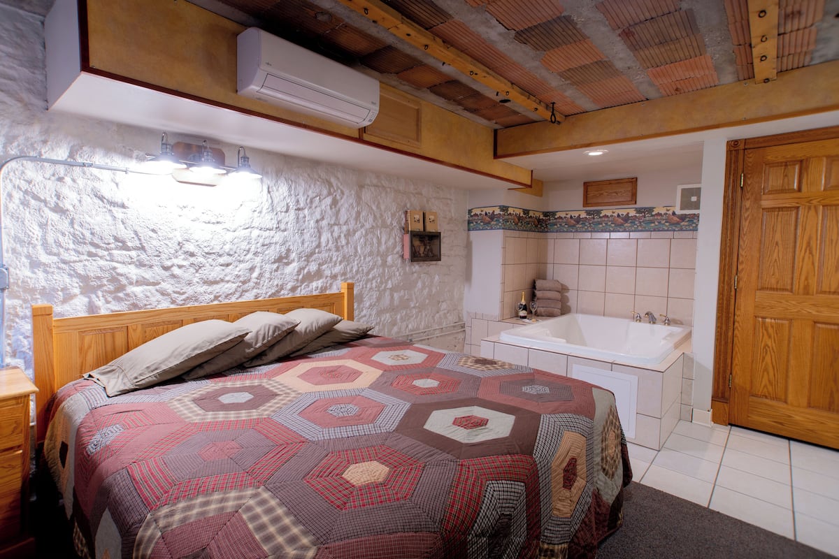 A bedroom with a large bed, a wooden headboard, a nightstand, and a built-in bench. There is a bathroom with a bathtub and a sink.