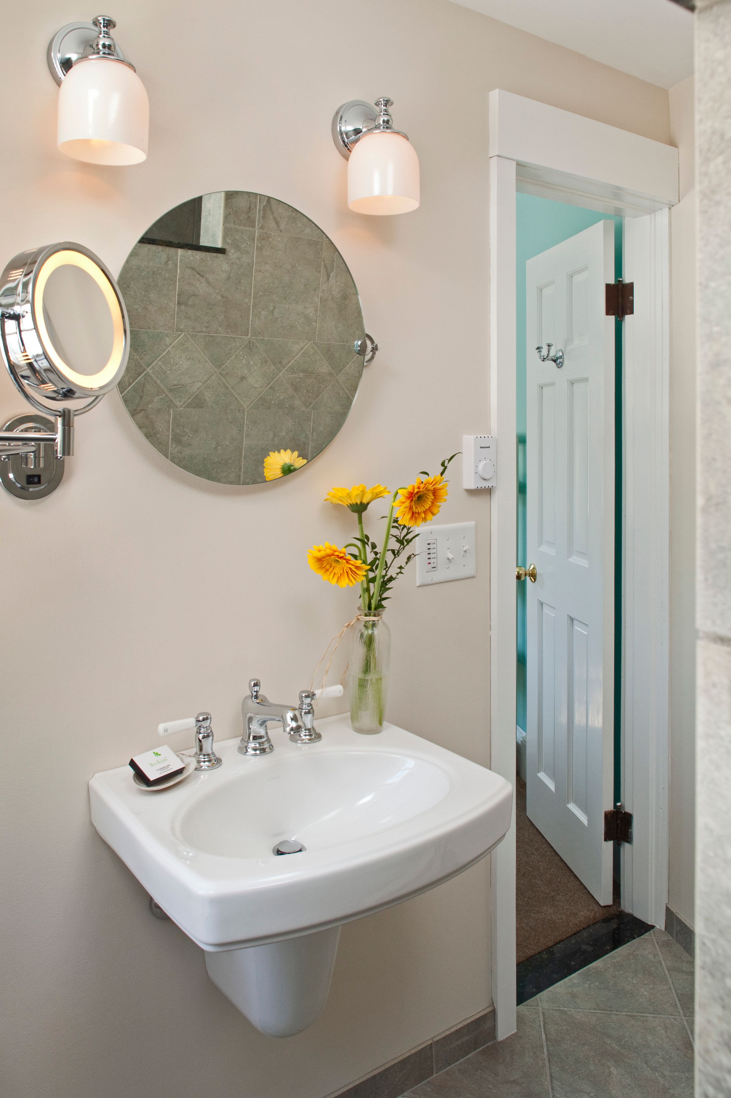 A bathroom with a white sink, a round mirror, and a vase with yellow flowers.