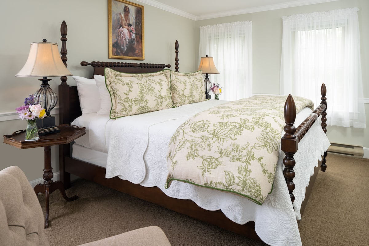 A neatly made bed with a floral bedspread, two bedside lamps, a nightstand with a vase of flowers, and a painting on the wall.