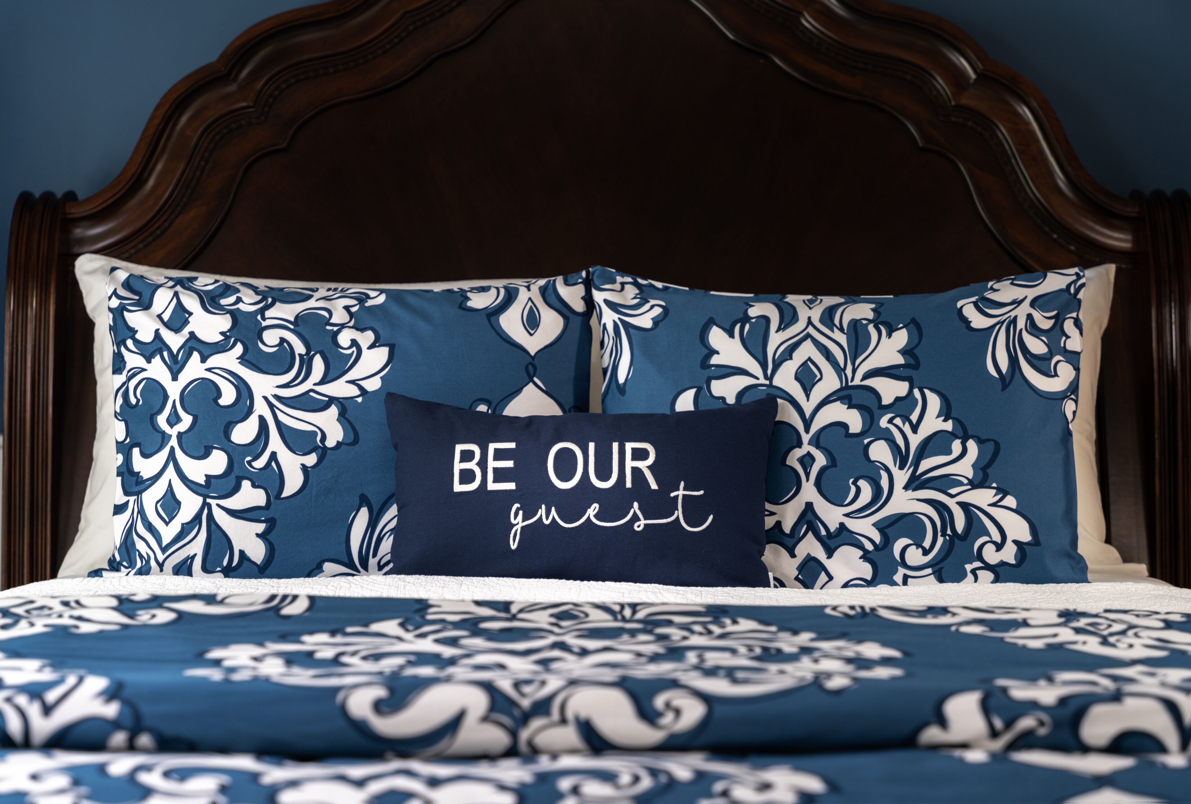 A bed with a dark wooden headboard, blue and white patterned bedding, and a pillow with the text "BE OUR guest".