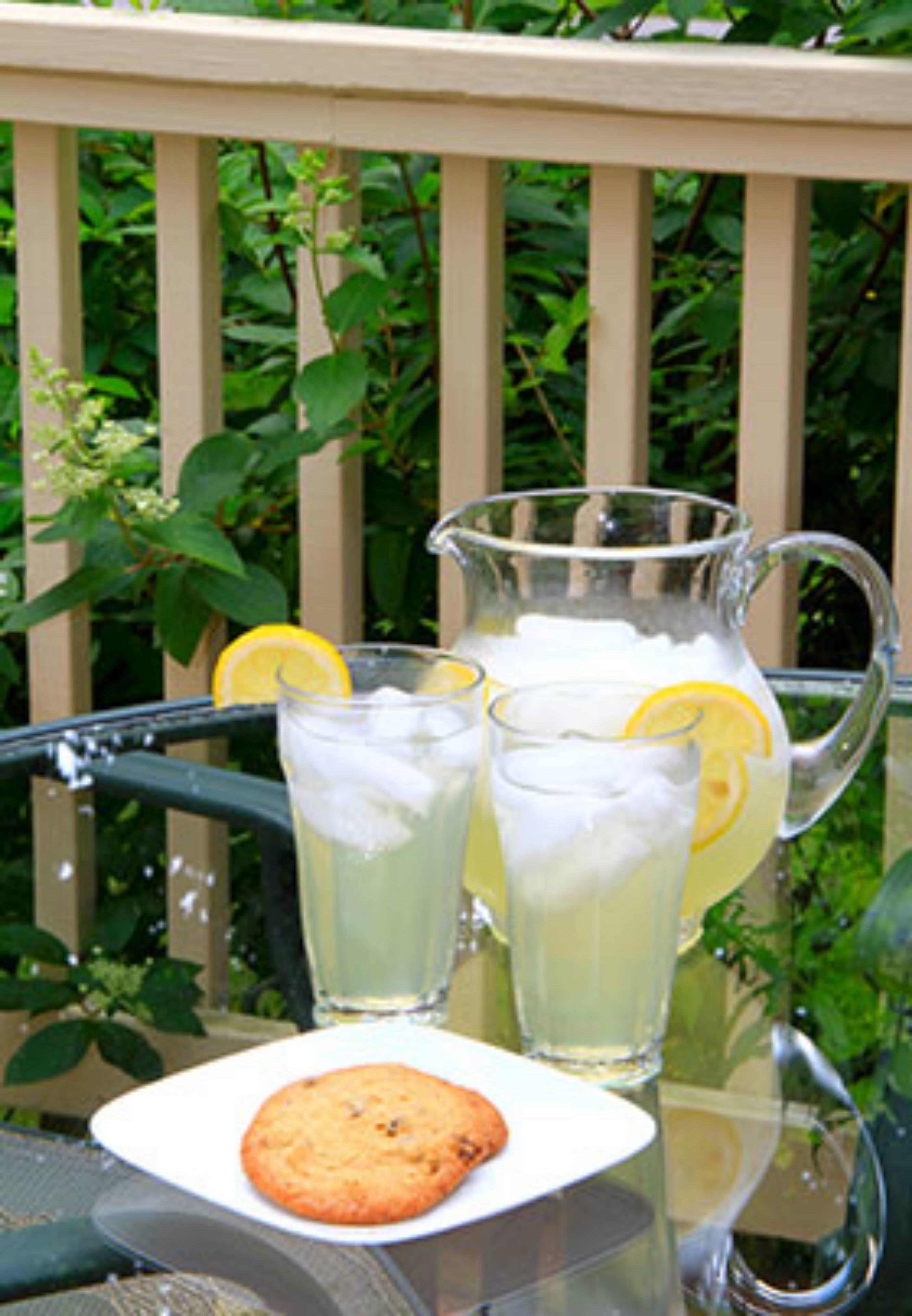 A glass pitcher and two glasses of lemonade with lemon slices on a table outdoors.